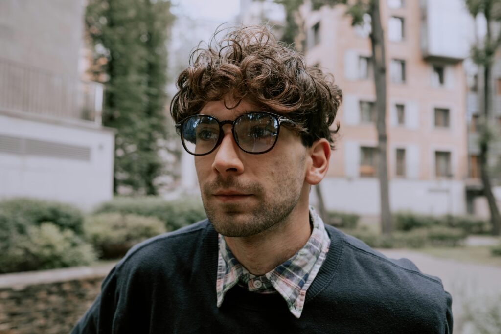 Portrait of a fashionable man with curly hair and glasses, standing outdoors.
